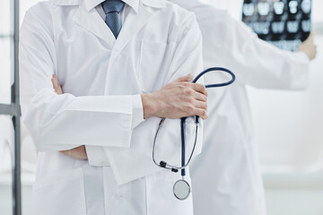 a doctor in a white coat and a stethoscope stands with his arms crossed in a hospital