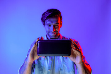 Smiling caucasian man holding and show VR headset with blurring background while standing with neon light. Smart teenager looking at digital goggles to enter virtual reality or metaverse. Deviation.