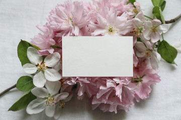 Closeup of blank business card, invitation mockup. Feminine scene with blurred pink blossoming Japanese cherry, apple tree branch. White linen table background. Flat lay, top view. Easter banner.