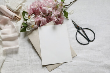 Wedding spring styled photo. Feminine scene mockup with pink blossoming Japanese cherry, apple tree branch. Blank paper greeting card, black scissors, white linen table background. Flat lay, top view. - obrazy, fototapety, plakaty