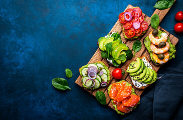Avocado toasts with salmon, shrimp, vegetables, spinach, capers and cream cheese, served on wooden board, blue table background, top view - 793287631