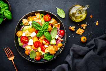 Summer vegetable salad with stale bread, tomatoes, cucumber, cheese, onion, olive oil, sea salt and green basil, black table background, top view