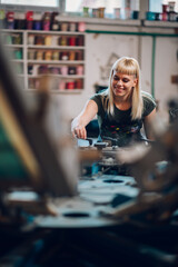 Fototapeta na wymiar Smiling female graphic worker adjusting screen printing press at factory