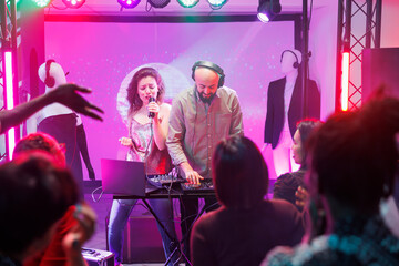 Woman singing and man dj mixing tracks while people partying on dancefloor in nightclub. People...