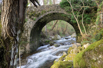 Old masonry stone bridge over the mighty Ambroz River and rocks with abundant moss