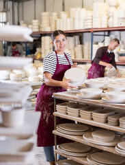 Positive young female artisan focused on stacking newly crafted ceramic plates on racks for even...