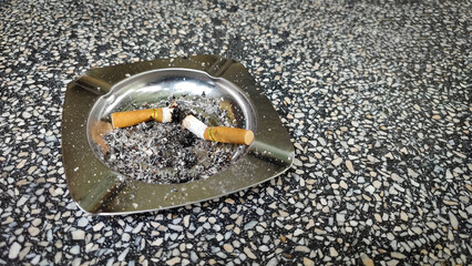 Silver ashtray, cigarette butts and ashes on a gray and white patterned surface, brown wooden table