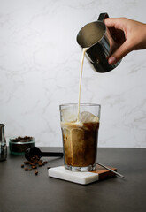 Pouring oat milk into cold ice coffee in a tall glass, grey marble table