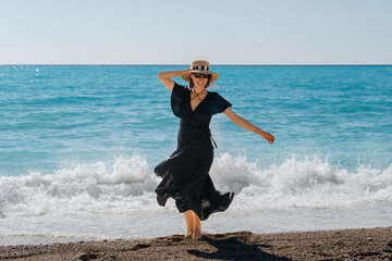 A joyous woman in a black dress and sun hat playfully splashes by the sea waves on a clear, sunny...