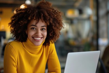 A woman with curly hair is smiling and sitting in front of a laptop. She is wearing a yellow sweater and she is happy