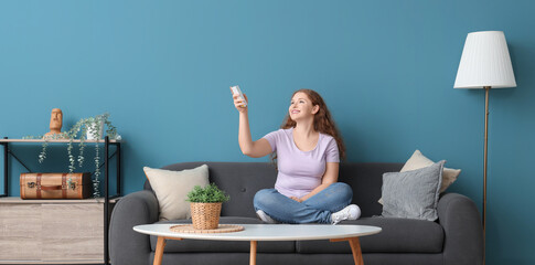 Young woman switching on air conditioner while sitting on sofa at home