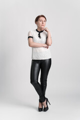 Confident businesswoman looking at camera with one hand raised to chin, standing legs crossed at ankle on white background. Female business person wearing blouse, leather trousers, high heeled shoes