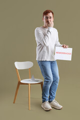 Young man holding paper with word HEMORRHOIDS and thumbtack on chair against green background