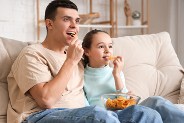 Happy father and his little daughter with nachos watching TV on sofa at home
