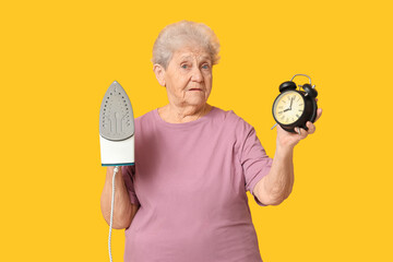 Senior woman with iron and alarm clock on yellow background