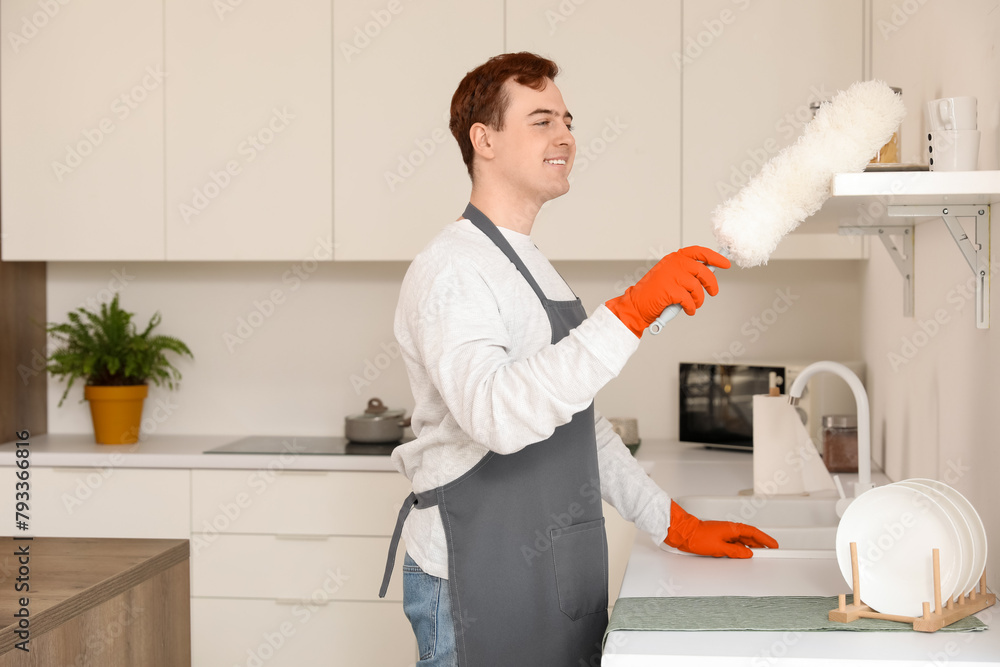 Wall mural Young man with pp-duster cleaning shelf in kitchen