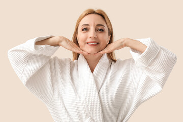 Beautiful happy mature woman in bathrobe doing face building exercise on beige background