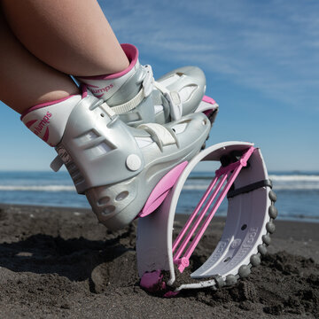 Closeup female legs in Kangoo Jumps shoes on black sandy beach while resting during fitness exercising. Low angle cropped shot of trendy pink sports jumping boots. Kamchatka, Russia - June 15, 2022