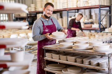 Experienced bearded artisan focused on stacking newly crafted ceramic plates on racks for even...