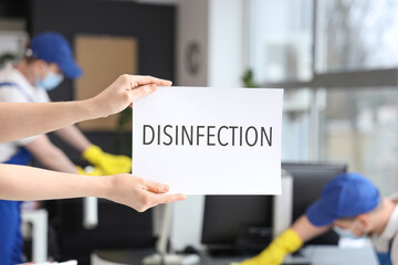 Worker of cleaning service holding paper with word DISINFECTION in office, closeup