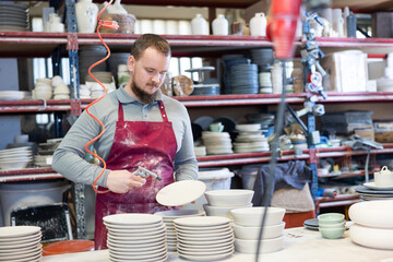 Skilled young bearded male artisan in maroon apron working in pottery workshop, dusting off...
