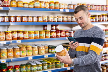 Adult male shopper with mobile phone choosing sauerkraut in grocery store