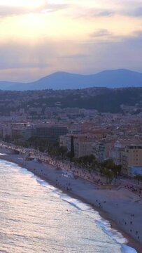 Picturesque scenic view of Nice, France on sunset. Mediterranean Sea waves surging on the beach, people are relaxing on the beach, cars driving the road. Camera vertical tilt. France