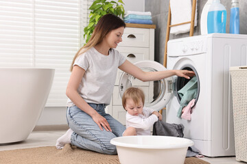Happy mother with her daughter putting baby clothes into washing machine in bathroom