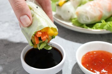 Woman dipping delicious spring roll into soy sauce at grey table, closeup