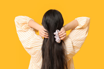 Woman tying hair with silk scrunchy on yellow background, back view