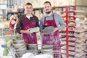 Two smiling male potters in stained maroon aprons holding handcrafted ceramic plates, conveying...