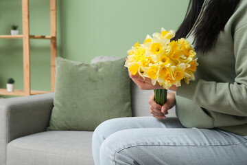 Woman holding bouquet with beautiful narcissus flowers and sitting on sofa in room