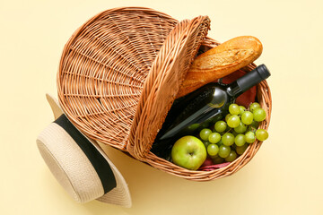 Fruits, baguette and wine in wicker basket near hat on beige background. Top view