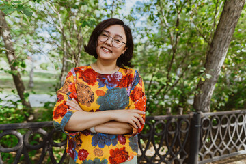 Smiling female with crossed arms standing in a lush park, exuding happiness Confident asian woman in glasses and bright blouse with gorgeous smile posing in summer park. Portrait looking at camera