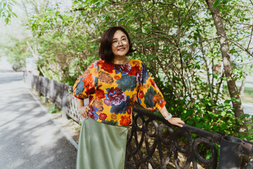A stylish Asian woman in a top with a floral pattern stands smiling at the railing of the park. Happy middle-aged woman feeling relaxed and content in a green park.