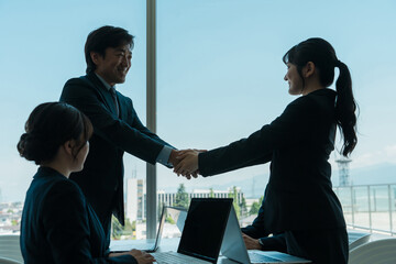 企業のオフィス・会議室で握手をして契約を結ぶ中高年ビジネスマンと若いビジネスウーマン（シルエット）
