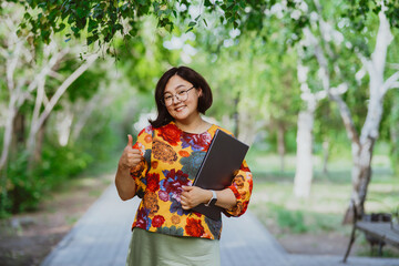 Smiling Asian woman with glasses and a laptop showing thumbs up in a sunny park. Portrait looking at camera. Happy businessman enjoying remote work among green trees outdoors