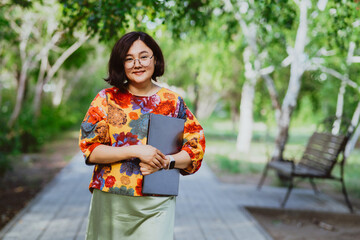Confident young professional woman with laptop in summer city park. Happy businessman enjoying a walk after working remotely among green trees outdoors