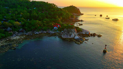 A tranquil tropical coastline basks in the amber glow of sunset, casting its golden spell upon the shimmering waters. Flight over the sea. Koh Tao, Thailand. Ocean background.
