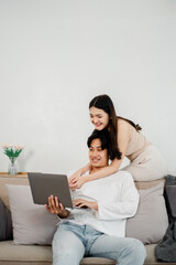 Smiling Asian couple is comfortably engaging with a laptop, showcasing a moment of shared discovery and enjoyment on their sofa.