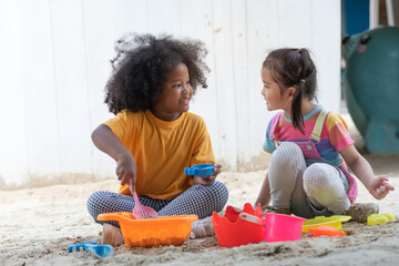 African little girl and her Asian friend play with sand in playground at park, summer outdoor activity for kids