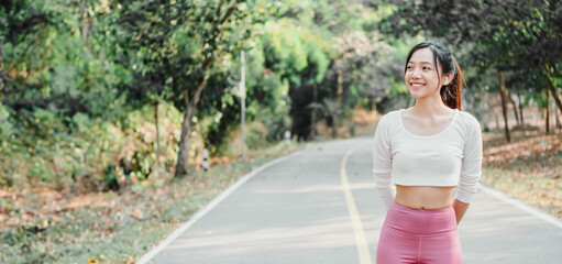 Young woman in a white crop top and pink leggings stands smiling on a sunlit park pathway with greenery in the background.