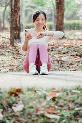 Woman seated on a park path looks at her fitness tracker while holding a smartphone, with scattered autumn leaves in the background.