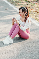 Seated woman in pink leggings and white sneakers uses her smartphone on a park road, with earphones in and trees in the background.