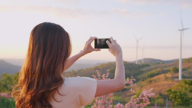 Asian woman using phone taking a photo outdoors in the wind turbine field. 