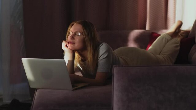 Relaxed adult woman lying with a laptop on sofa, the setting sun's rays fall on her face. Female blonde having relax and indoor technology leisure activity with wireless connection.