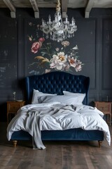 A dark blue velvet bed with a navy headboard, white sheet and pillows in the center of a large room with dark gray walls and a vintage chandelier