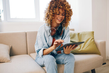 Happy Woman Enjoying Leisure Time at Home: Holding Tablet PC, Listening to Music with Headphones, and Smiling on Cozy Couch.