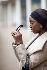 An African American woman is depicted examining a pair of sunglasses outdoors. She's dressed in a light beige coat with a black belt and headband, exuding a sense of casual urban fashion. The shallow