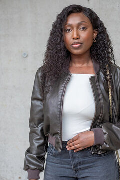 A poised African American woman stands against a concrete wall, exuding confidence. Her curly black hair frames her face, complemented by subtle makeup. She's clad in a modern ensemble featuring a